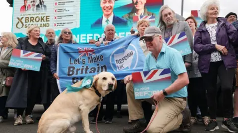 Getty Images A group of people holding Reform signs and banners line up behind a large flag. There man with a Reform sign and a labrador dog with a Reform rosette in the foreground, and a large Reform billboard in the background. 