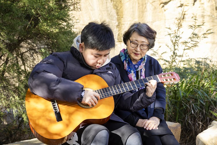 A boy playing a guitar outdoors alongside a woman.