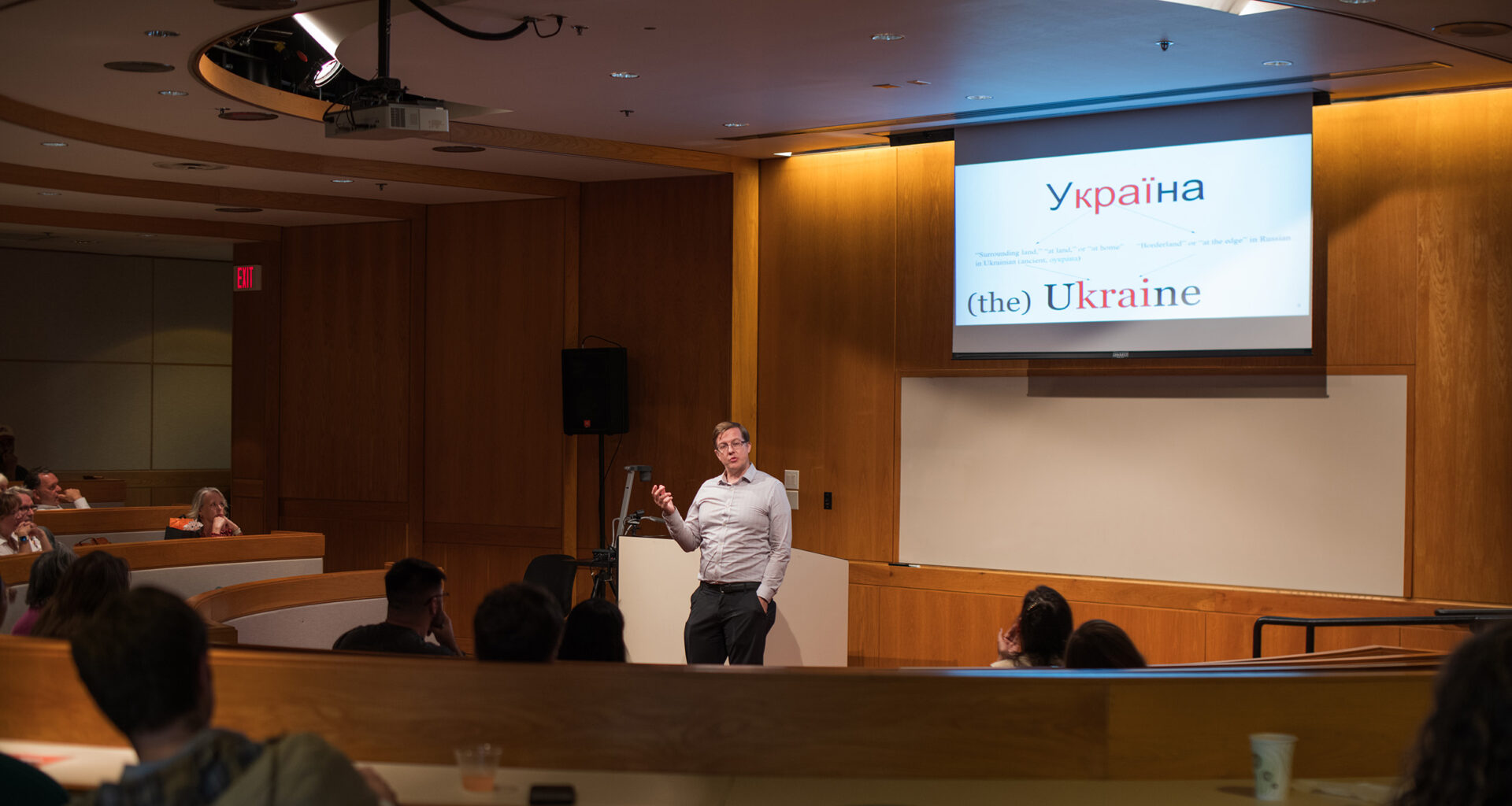 A presenter stands at a podium in a lecture hall, addressing an audience seated in curved rows. Behind the presenter, a large screen displays a slide with the word "Україна" (Ukraine) and explanatory text in English about the correct usage of "Ukraine" versus "the Ukraine.