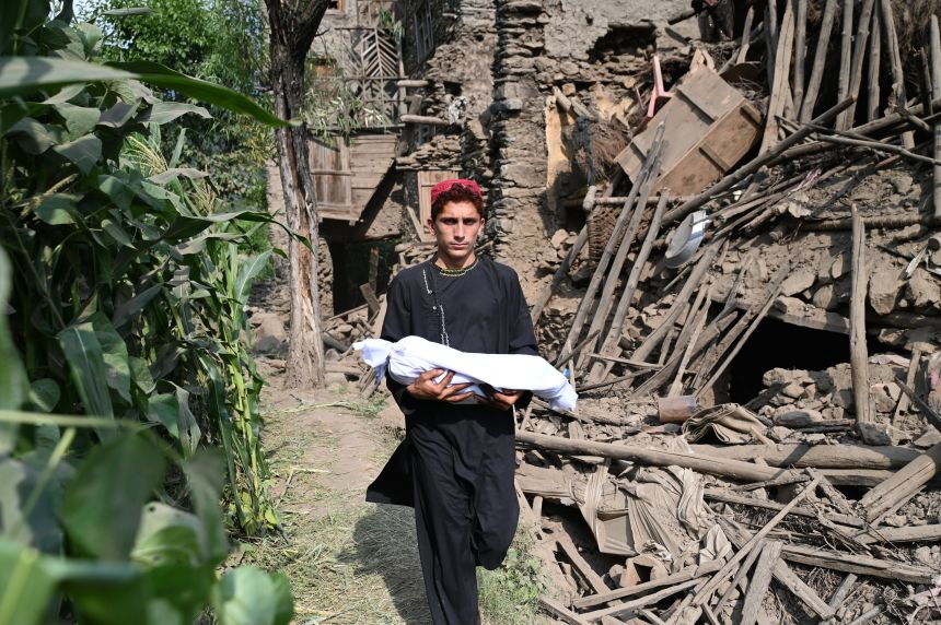 An Afghan boy carries a baby's body in a shroud as he walks by a damaged house following earthquakes in the Mazar Dara village of Nurgal, a district of the Kunar Province, in Eastern Afghanistan, on September 1, 2025.