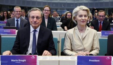 Mario Draghi and Ursula von der Leyen sit side by side at a conference table in Brussels before delivering a keynote address, with attendees in the background