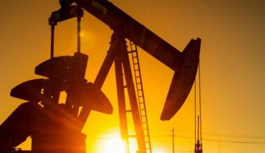 A pump jack silhouetted against a bright sunset, with oil extraction equipment visible in the foreground.