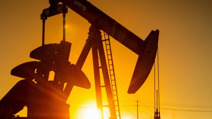 A pump jack silhouetted against a bright sunset, with oil extraction equipment visible in the foreground.