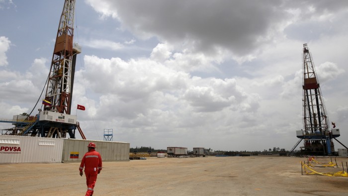 An oilfield worker in red coveralls walks near two drilling rigs at a PDVSA oil well in Venezuela’s Orinoco belt.