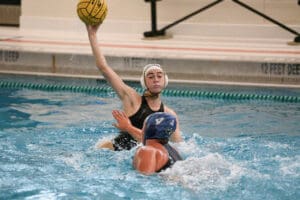 Water polo player in black ready to throw the ball against an opponent in blue cap, indoor pool action.