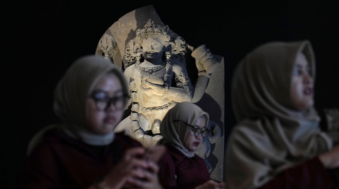 Three women are visible in the blurred foreground, with a stone statue in the background. The status is in focus and lit by a spotlight.