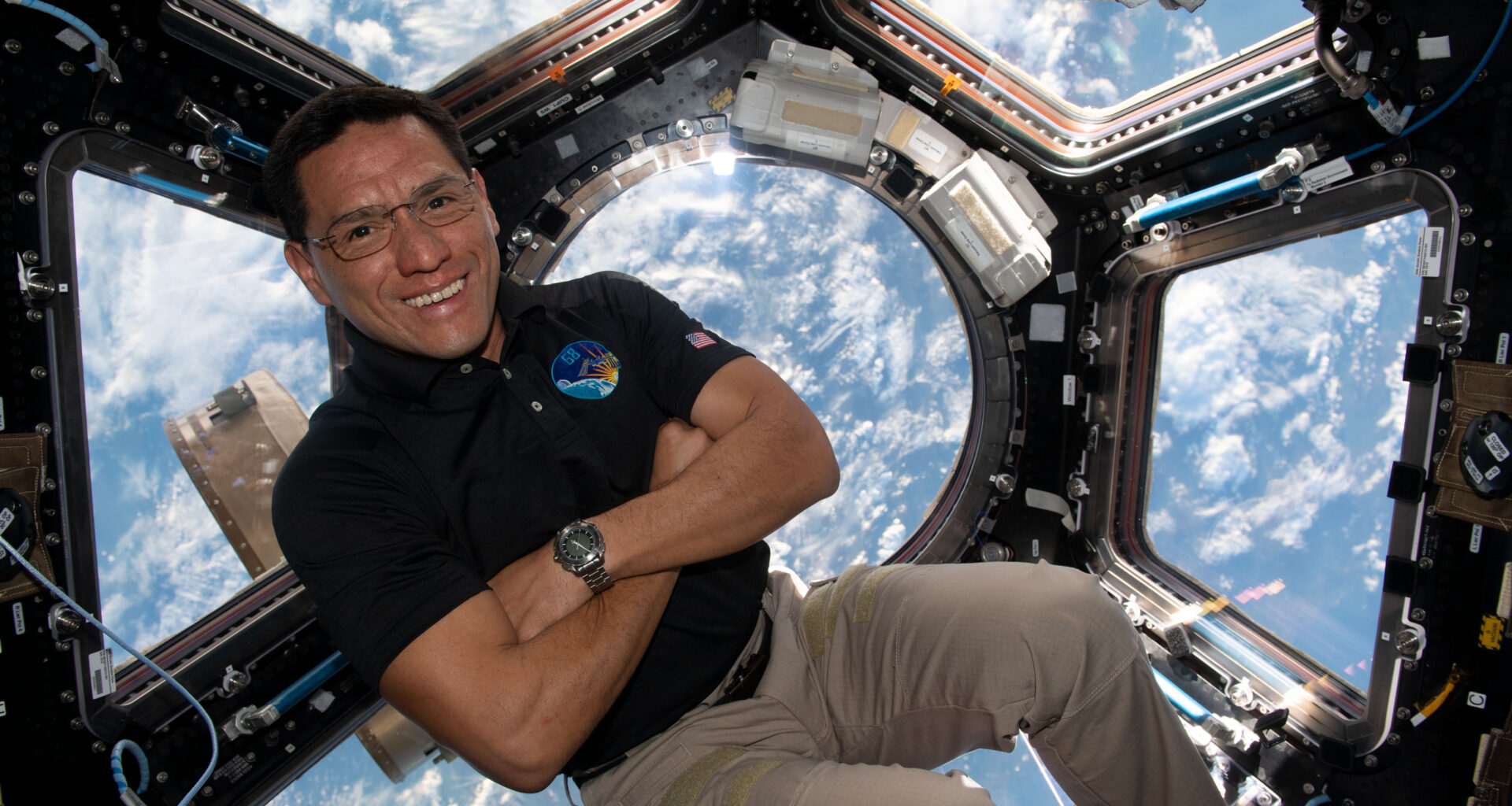 A man in a dark polo shirt smiles at the camera with his arms crossed. He is in the cupola of the International Space Station, an area with multiple windows through which Earth and space can be seen. Earth's clouds can be seen through the windows behind him.