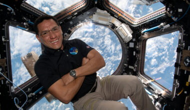 A man in a dark polo shirt smiles at the camera with his arms crossed. He is in the cupola of the International Space Station, an area with multiple windows through which Earth and space can be seen. Earth's clouds can be seen through the windows behind him.