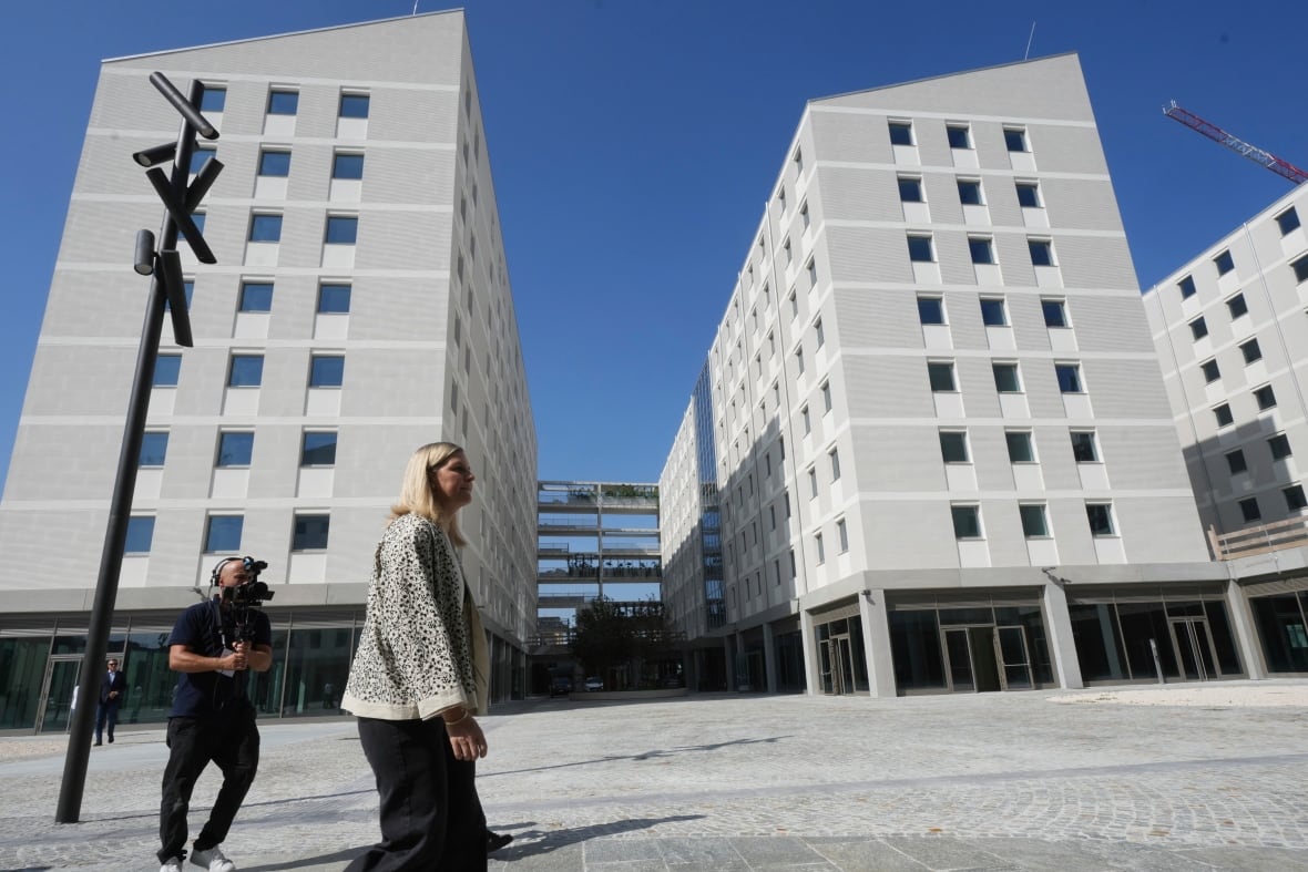 Two people walk past tall, white buildings.