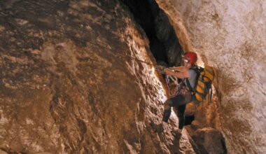 Hazel Barton, wearing a backpack and head lamp, crosses a sheer face of cave wall holding onto a rope with her boots braced against the rock.