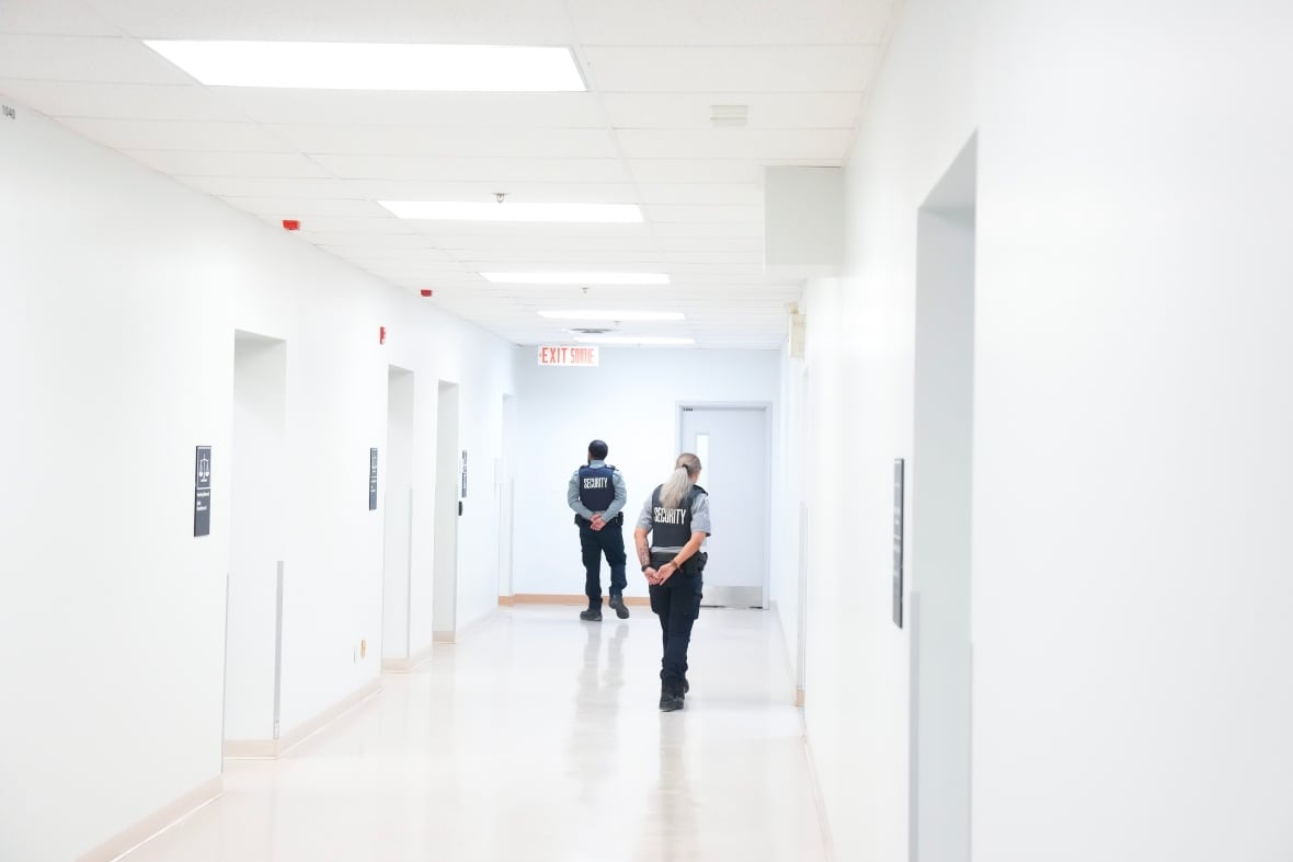 The backs of two security guards walking down a white hallway of an immigration holding centre in Toronto.