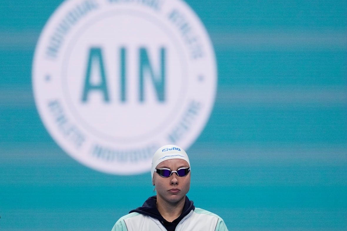 A swimmer wearing goggles and a swim cap stands behind a sign that says "AIN."