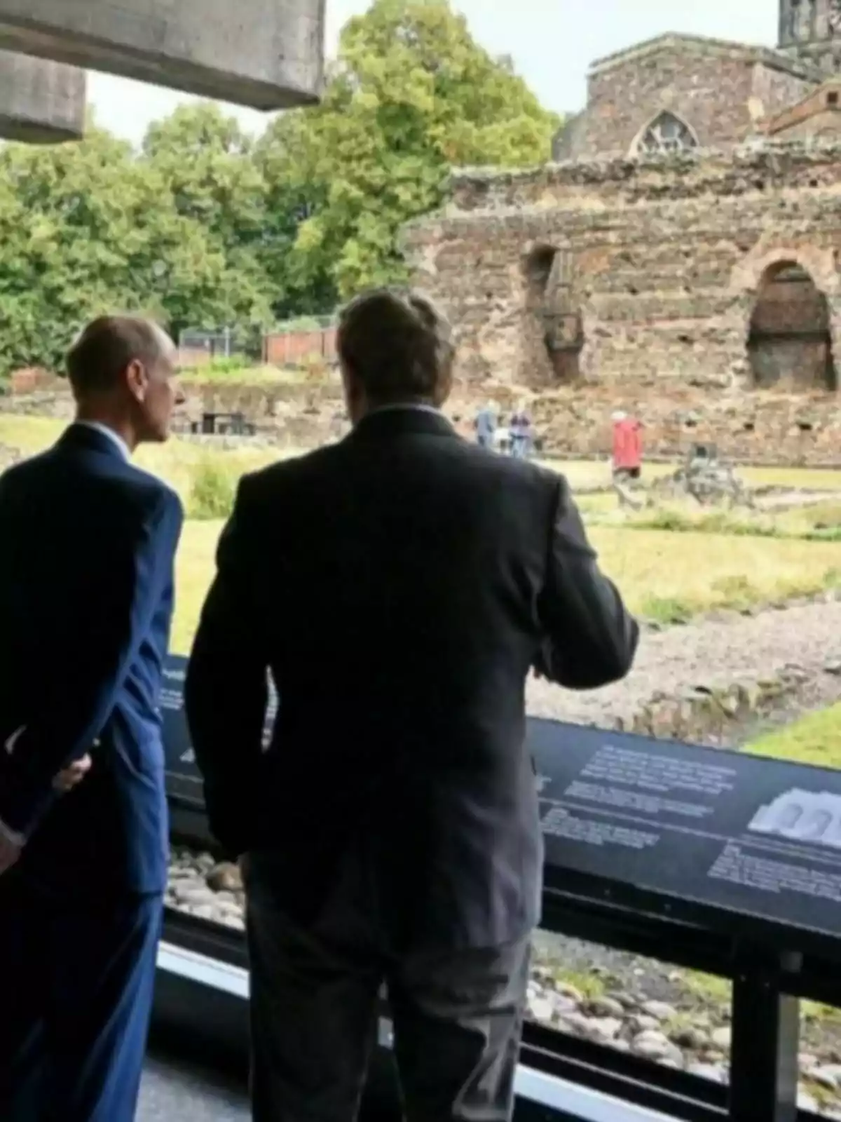 Two men with their backs turned observe ancient ruins through a large window, surrounded by trees and informational panels.