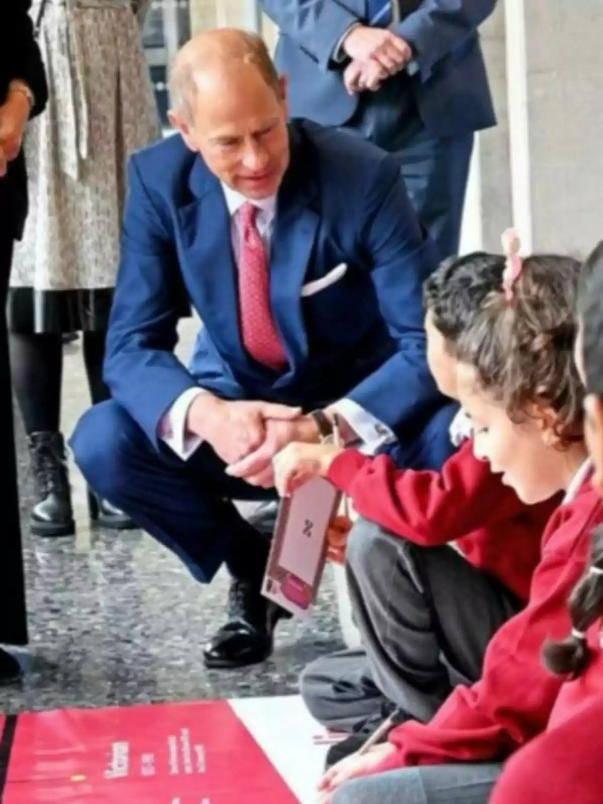Prince Edward in a blue suit interacting with a girl holding a notebook while other children watch