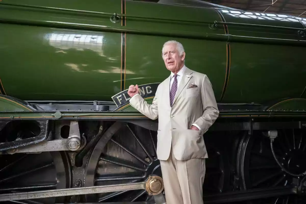 Charles III with gray hair and a beige suit posing next to an old green locomotive.