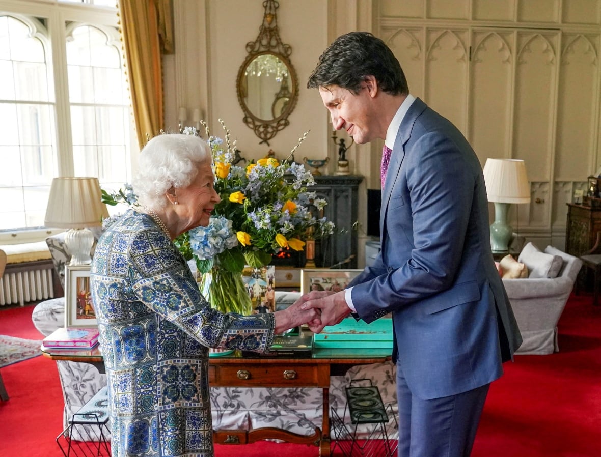 Two people shake hands in a large sitting room in front of a large bouquet of flowers.