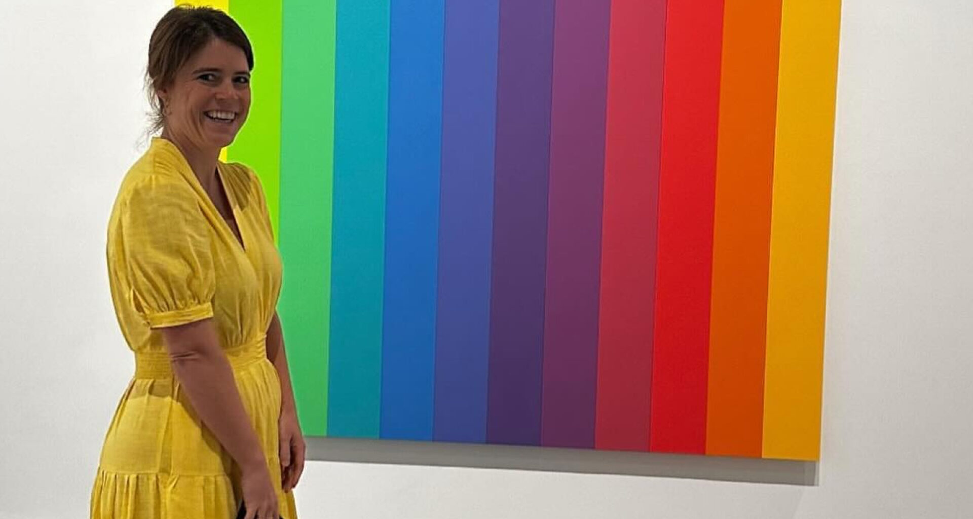 Princess Eugenie wearing a yellow dress standing in front of a rainbow painting