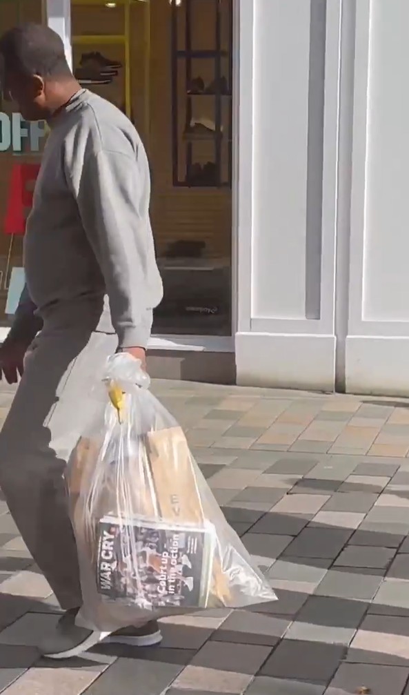 A man in a grey tracksuit carrying a clear bag with newspapers, one titled "WAR CRY," and other items.