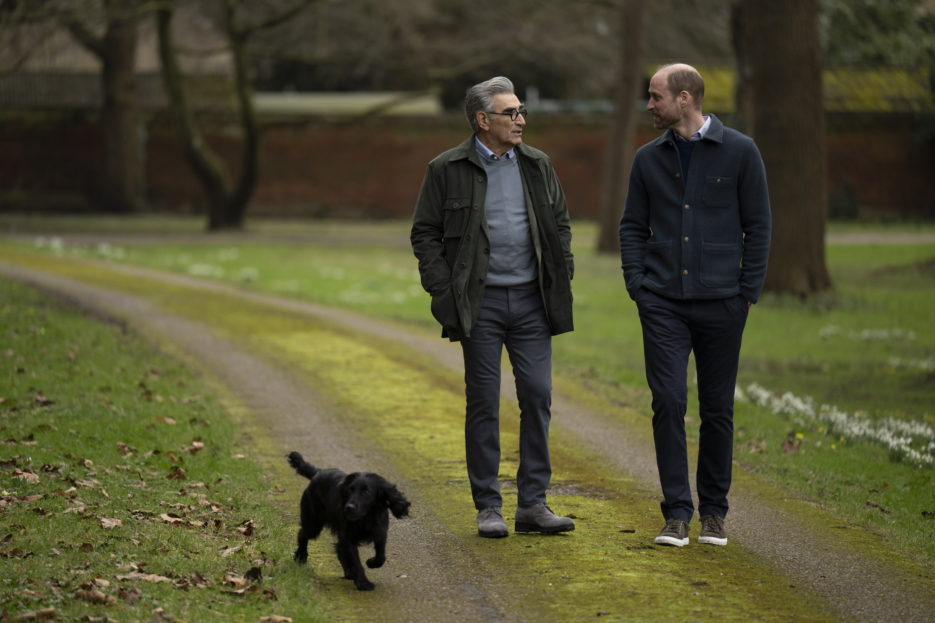 Eugene Levy and Prince William walking outside at Windsor Castle with a black dog.