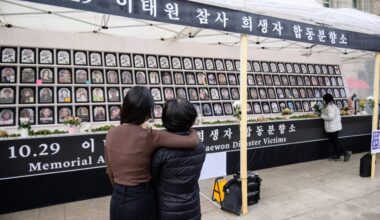 A woman with dark hair wearing a brown shirt wraps her arm around another woman with dark hair wearing a black coat as they look at a large memorial display of victims’ portraits. A black banner with white Korean letters appears above the display.