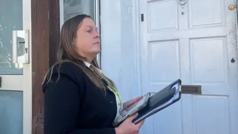 BBC A woman with a clipboard and lanyard outside a white-painted wooden door.