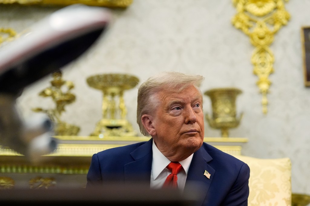 A person in a navy suit and red tie sits indoors against a backdrop of ornate gold decorations, looking to the side.