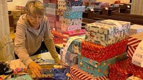 BBC A woman in a grey zip up jumper and white top. She is reaching over the table for an elastic band in a black box filled with elastic bands. In front of her and to the right of her are piles of shoeboxes on tables wrapped in wrapping paper. To the right are church pews also filled with wrapped shoeboxes. 