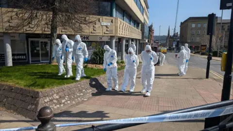 Alex Moss/BBC A number of forensic officers in white suits walk through Huddersfield in a crime scene, there is police tape in the foreground. 