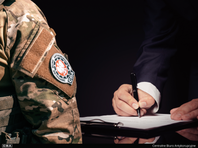 Funkcjonariusz CBA, za nim mężczyzna podpisuje pismo.