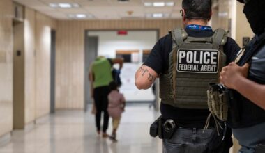 A photo shows the back of a light-skinned officer wearing glasses, a black T-shirt, gray jeans and a vest with the words "police federal agent." One police officer stands next to them. A person and a child walk down a hallway standing in the background.