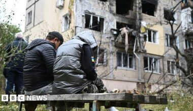 Two people sit on a bench against the backdrop of a destroyed building