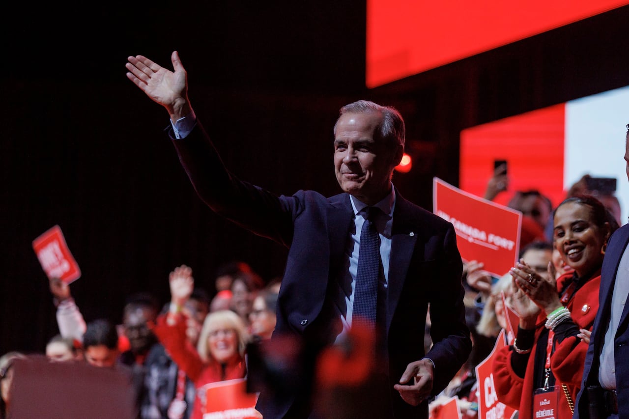 A smiling grey-haired man in a dark blue business suit waves as two dozen happy people dressed in red applaud behind him.