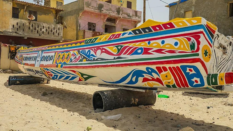 A colourful fishing canoe seen from an angle at the back. It is sitting on rollers on the beach. Some buildings at the beachside can be seen in the background.