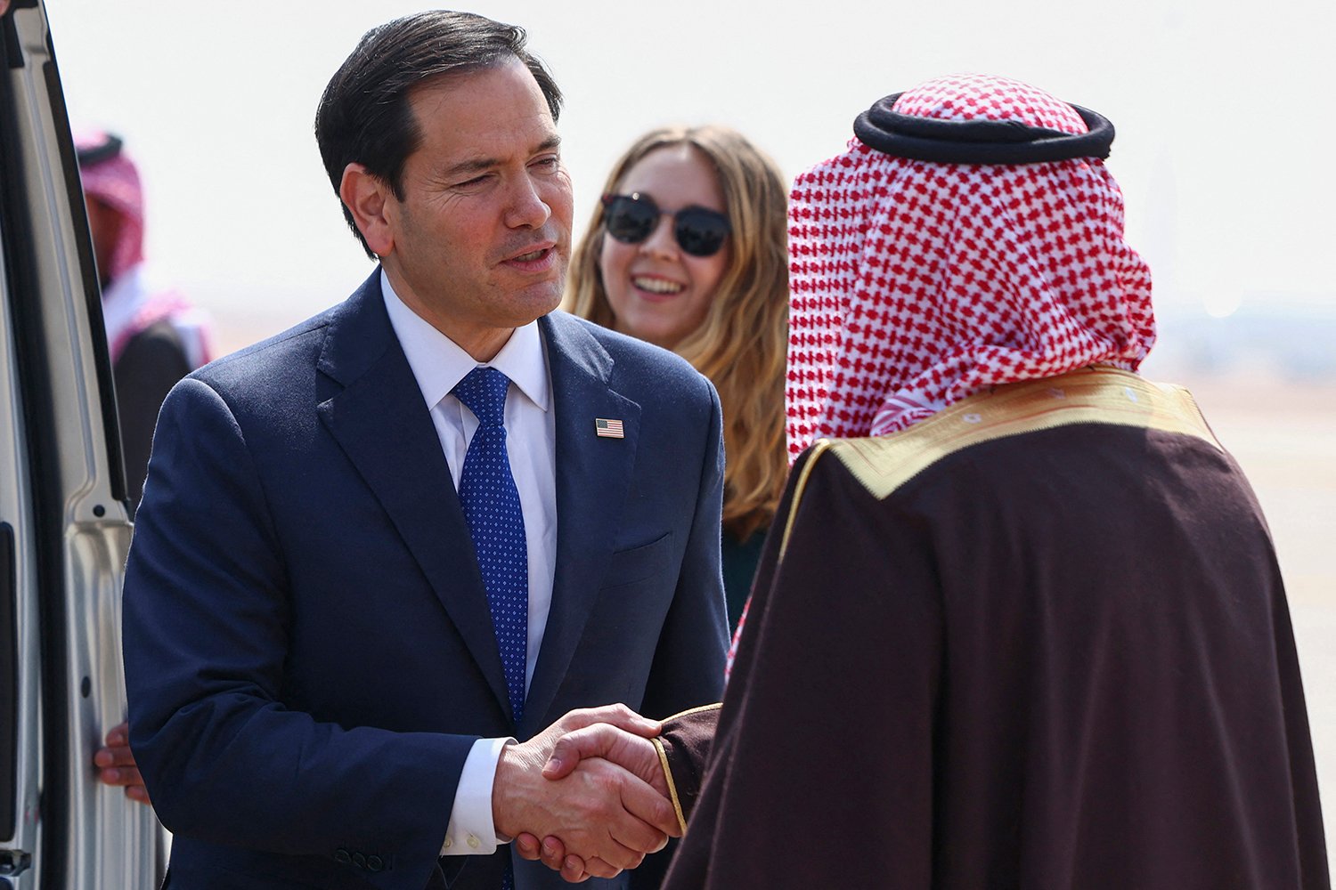 Marco Rubio shakes hands with Abdulmajeed al-Smari as they stand together outdoors on a sunny day. Other people mill about behind them.