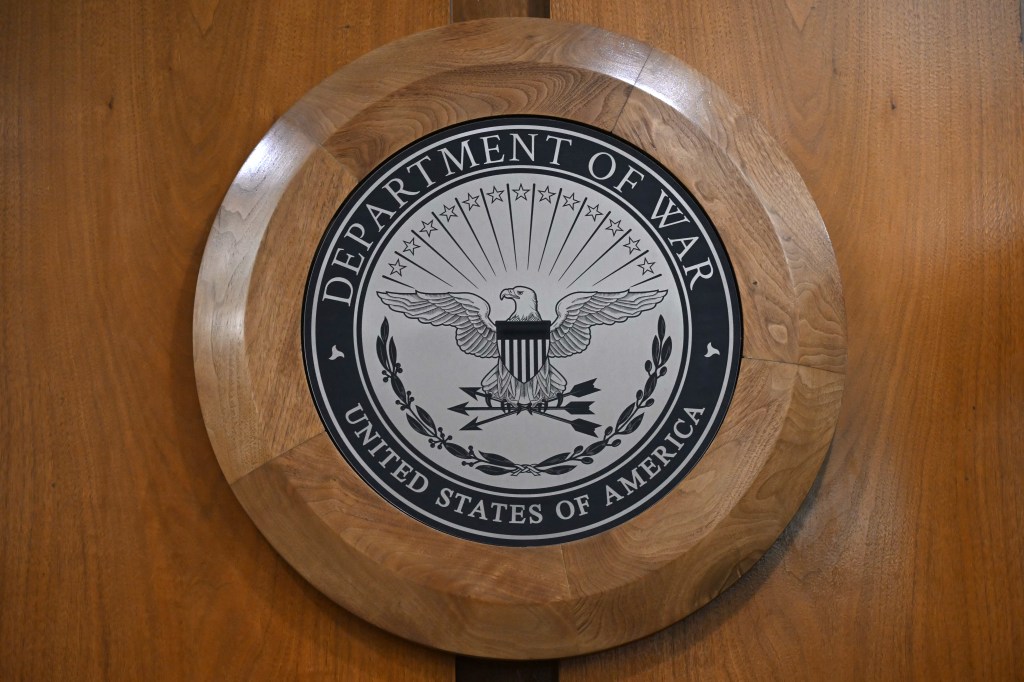The Department of War logo, featuring an eagle and arrows, set against a wooden background.