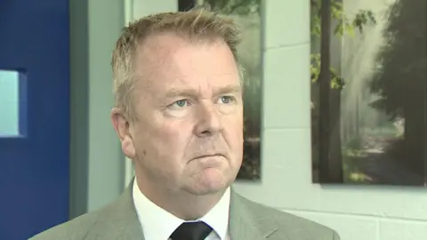 Kevin Shortall. He has short brown-ish hair, wearing a grey blazer, white shirt and black tie.