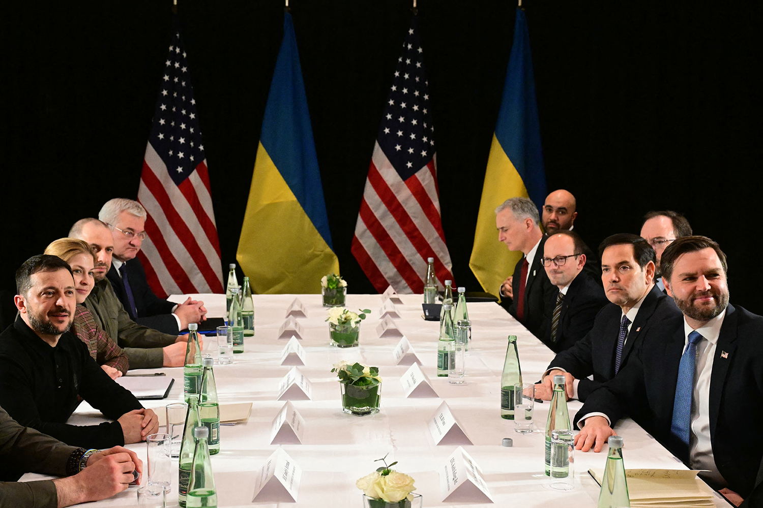 United States and Ukrainian flags hang behind a table surrounded by a seated group that includes J.D. Vance, Marco Rubio, and Volodymyr Zelensky. The table is covered in a white tablecloth, and each attendee has a name card, green water bottle, and glass in front of them.