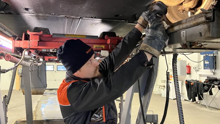 A man with a moustache, black knit cap and orange and grey coveralls works under a car up on a lift.
