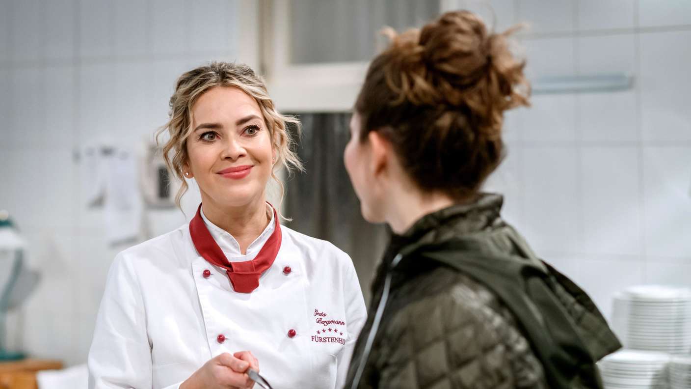 Greta aus „Sturm der Liebe“ steht links Fanny gegenüber und schaut sie lächelnd an. 