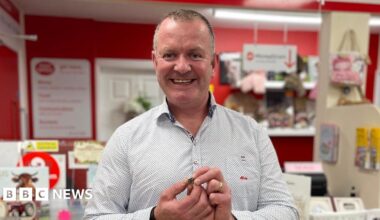Sean Farry. He has short shark hair, wearing a light blue shirt. He is smiling and holding the 5p coin in his hands.