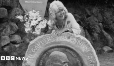 A black and white photo shows a woman with shoulder length grey hair leaning over a rock which is engraved with the words 'Darwin gardens millennium green'. She is holding a bunch of flowers and smiling.