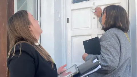 Two women with clipboards knock at a white painted wooden door