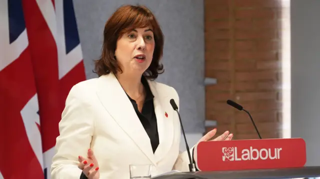 Lucy Powell in white blazer and black top speaks behind a podium with two black mics in front of her and Union flags behind her