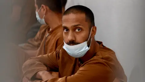 Goktay Koraltan/BBC Hamza Parvez stares whilst wearing a mask and a dark brown top. 