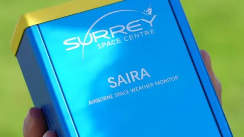 Tamsin Melville/BBC A blue box has the words Surrey Space Centre Saira Airborne Space Weather Monitor on it. It is held on someone's hands, to give you an idea of its size.