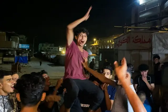 Un grupo de hombres jóvenes palestinos están en círculo con caras de júbilo y sostienen a uno de ellos elevado, sobre los hombros de uno otro. El que está elevado tiene su brazo levantado, con una gran expresión de júbilo. Todos visten ropa casual, camisetas y pantalón deportivo. Es de noche.