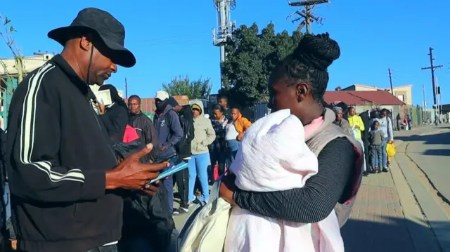 Side view of man wey wear black tracksuit top and dark sunhat as e face woman wey hold baby. Di man dey check some documents.