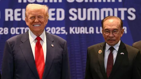 AFP via Getty Images Donald Trump and Anwar Ibrahim are wearing dark suits and standing in front of a dark blue background. Trump is grinning.