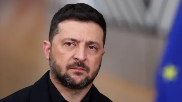 A close up image of Zelensky in front of a blurred European Union flag. He's wearing a black shirt and has a grim expression on