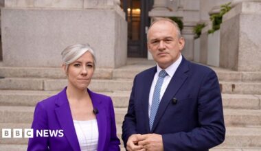 Sir Ed Davey and Daisy Cooper stand outside the Bank of England. Ed Davey wears a dark blue suit, white shirt, and blue patterned tie; Daisy Cooper wears a purple blazer over a white top. Behind them are stone steps and the grand entrance of the Bank of England